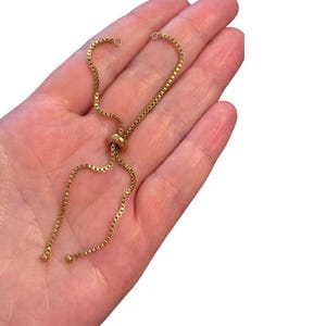 May include: A gold-toned box chain bracelet featuring an adjustable sliding bead mechanism. The chain consists of small, square-linked segments with a polished finish. Two small metal beads are attached to the ends of the chain strands.