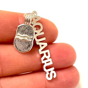 May include: Silver charm with a wave design and the word "Aquarius" on a separate charm. The charms are hanging from a silver loop.