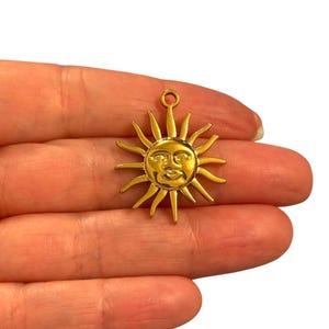 May include: A gold-colored sun charm with a smiling face and radiating rays. The charm has a loop at the top for attaching to a necklace or bracelet. The sun charm is held in a person's hand.