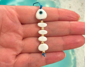 May include: A white and gold beaded charm with a blue string. The charm features a white eye-shaped bead with a blue center, followed by a series of white oval beads and gold beads. The charm is held in a hand.