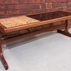 May include: A mid-century coffee table with a rectangular design. It has a wooden frame, a tiled section with a patterned design in orange, brown, and cream, and a glass top. The table's legs are curved. The dimensions are approximately 120 cm long.