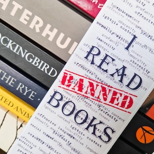 Könnte beinhalten: Ein Lesezeichen mit dem Text "I READ BANNED BOOKS" in Rot und Schwarz gestempelt, mit einer Liste verwandter Wörter. Das Lesezeichen befindet sich zwischen den Buchrücken mehrerer Bücher, darunter "To Kill a Mockingbird".