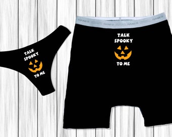 Op de afbeelding: Zwarte ondergoed- en boxershortset met een Halloween-thema. Beide stukken hebben een pompoenafbeelding en de tekst "TALK SPOOKY TO ME" in het wit. De achtergrond is een wit houten oppervlak.