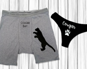 Op de afbeelding: Een paar grijze boxershorts met de tekst "Cougar Bait" en een zwart silhouet van een poema. Een zwarte string met de tekst "Cougar" en een pootafdruk.