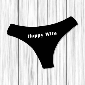 以下が含まれることがあります： 「Happy Wife」の白い文字が入った黒い下着。白い木製の背景に下着が配置されています。