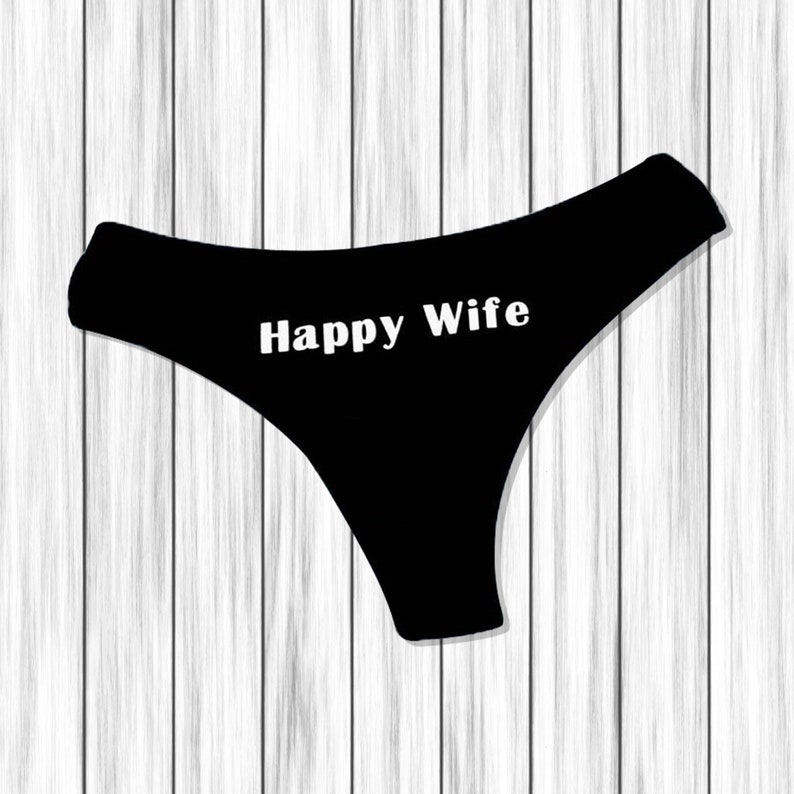 以下が含まれることがあります： 「Happy Wife」の白い文字が入った黒い下着。白い木製の背景に下着が配置されています。