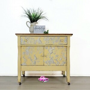 May include: A yellow painted vintage side cabinet with two doors and a drawer. The cabinet has a floral pattern with birds and is on wheels. The cabinet has a wooden top and is decorated with a sign that says "family".