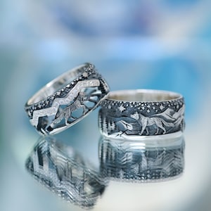 Puede incluir: Dos anillos de plata con grabados detallados de lobos y montañas. Un anillo presenta una fila de pequeñas piedras transparentes.