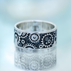 May include: A silver ring with a black gear design. The ring is reflecting the light, creating a mirror image of the design.