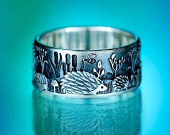 May include: A silver ring with a detailed design of hedgehogs, plants, and a bee. The ring is on a blue background.