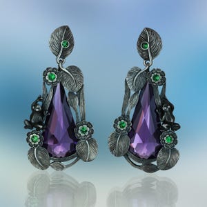 May include: A pair of silver-toned earrings featuring large, faceted purple teardrop-shaped gems. The earrings are adorned with detailed leaf and flower accents, small green gemstones, and a small frog. The design is intricate and nature-inspired.