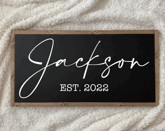 May include: Black wooden sign with a white cursive inscription that reads "Jackson EST. 2022".