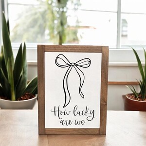May include: A framed wooden sign with a white background features a black line drawing of a bow. Below the bow, the words "How lucky are we" are written in a cursive font. The frame is a medium brown color.
