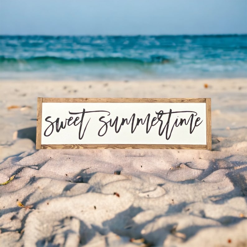 May include: A rectangular wooden sign with the words "sweet summertime" in black script lettering. The sign has a white background and a brown wooden frame. The sign is resting on a sandy beach with the ocean in the background.