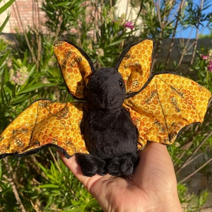 May include: A black plush bat with yellow and brown wings featuring a honeycomb pattern and bees. The wings are spread out and the bat is sitting on a hand.