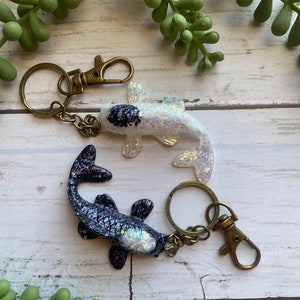May include: Two iridescent koi fish keychains with gold tone key rings and clasps. The fish are made of resin and have a glittery finish. One fish is white with black markings and the other is black with white markings.