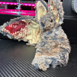 May include: A plush toy bat with gray and brown fur, featuring wings with a map design and the text "Marauder's Map". The bat has a small black nose and is set against a dark background.