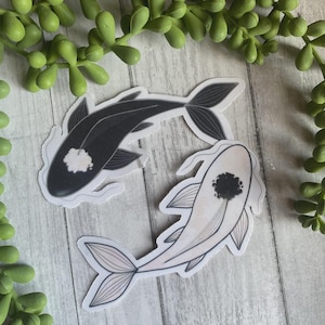 May include: Two koi fish stickers in a yin-yang arrangement. One fish is black with a white spot, the other is white with a black spot. The stickers have detailed fin and tail designs. Green succulent plants are in the background.
