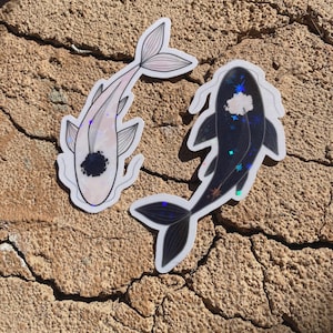 May include: Two koi fish stickers. One is light pink with a black spot and the other is dark blue with a white cloud and star design. Both stickers have a holographic shimmer and are on a textured, brown surface.