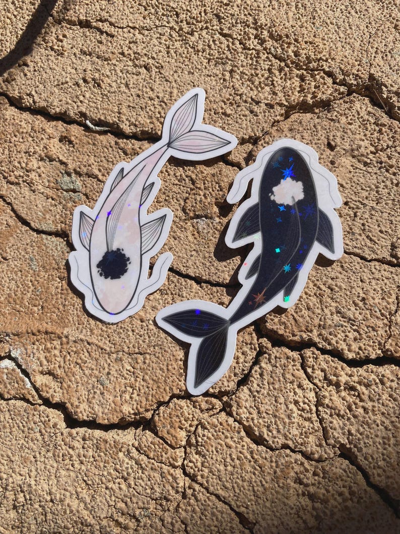 May include: Two koi fish stickers. One is light pink with a black spot and the other is dark blue with a white cloud and star design. Both stickers have a holographic shimmer and are on a textured, brown surface.