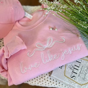 May include: A pale pink sweatshirt embroidered with the words "love like jesus" in white script. A matching pink baseball cap is next to the sweatshirt, along with pearl earrings and a book titled "Be Still".