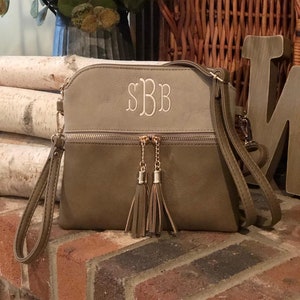 May include: A taupe-colored crossbody bag with a zipper closure and two tassels. The bag has a light brown top flap with the monogram "SBB" embroidered in white.