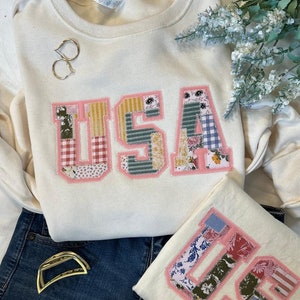 May include: A cream-colored sweatshirt with a large patchwork "USA" design in pink, yellow, blue, and green. The sweatshirt is laid out on a pair of blue jeans.