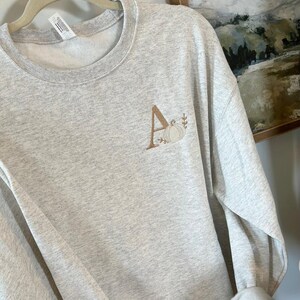 May include: A grey sweatshirt with a brown embroidered letter A and a pumpkin design.
