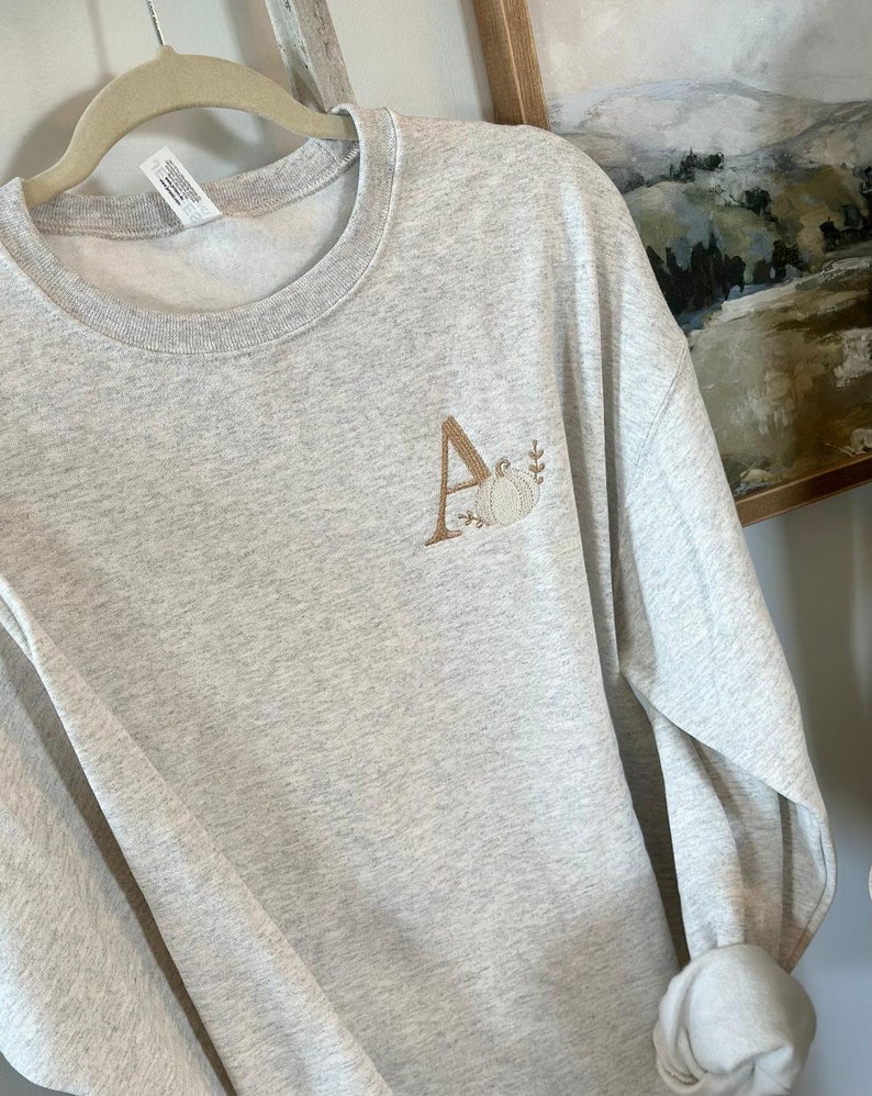 May include: A grey sweatshirt with a brown embroidered letter A and a pumpkin design.