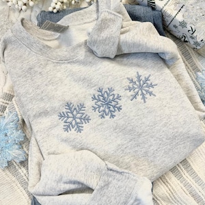 May include: A gray sweatshirt with three embroidered snowflakes in blue. The sweatshirt is folded and lying on a white and gray striped surface.