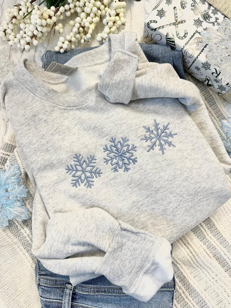 May include: A gray sweatshirt with three embroidered snowflakes in blue. The sweatshirt is folded and lying on a white and gray striped surface.