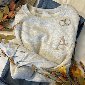 May include: A gray sweatshirt with a gold embroidered letter "A" and a pumpkin design. The sweatshirt is folded and laid on a white surface with a pair of gold hoop earrings and a sprig of dried flowers.