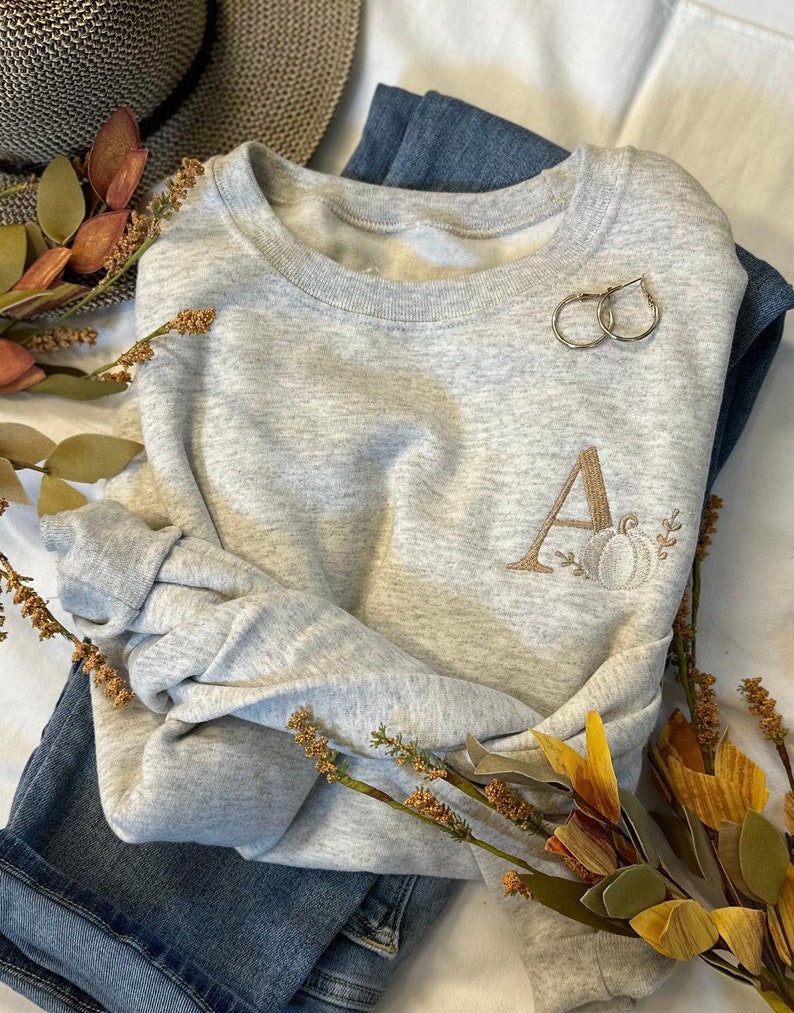May include: A grey sweatshirt with a gold embroidered letter "A" and a pumpkin design. The sweatshirt is folded and laid on a white surface with a pair of gold hoop earrings and a sprig of dried flowers.