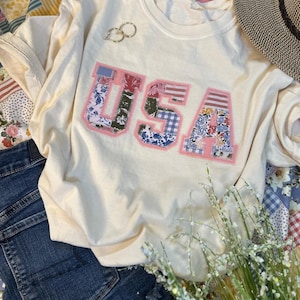 May include: A white t-shirt with a patchwork "USA" design in pink, blue, and green. The shirt is laid out on a patchwork quilt with a straw hat and a bouquet of white flowers.