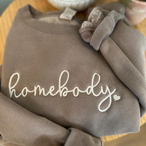 Peut inclure: Un sweat-shirt marron avec le texte "homebody" brod&eacute; en blanc avec un petit c&oelig;ur.