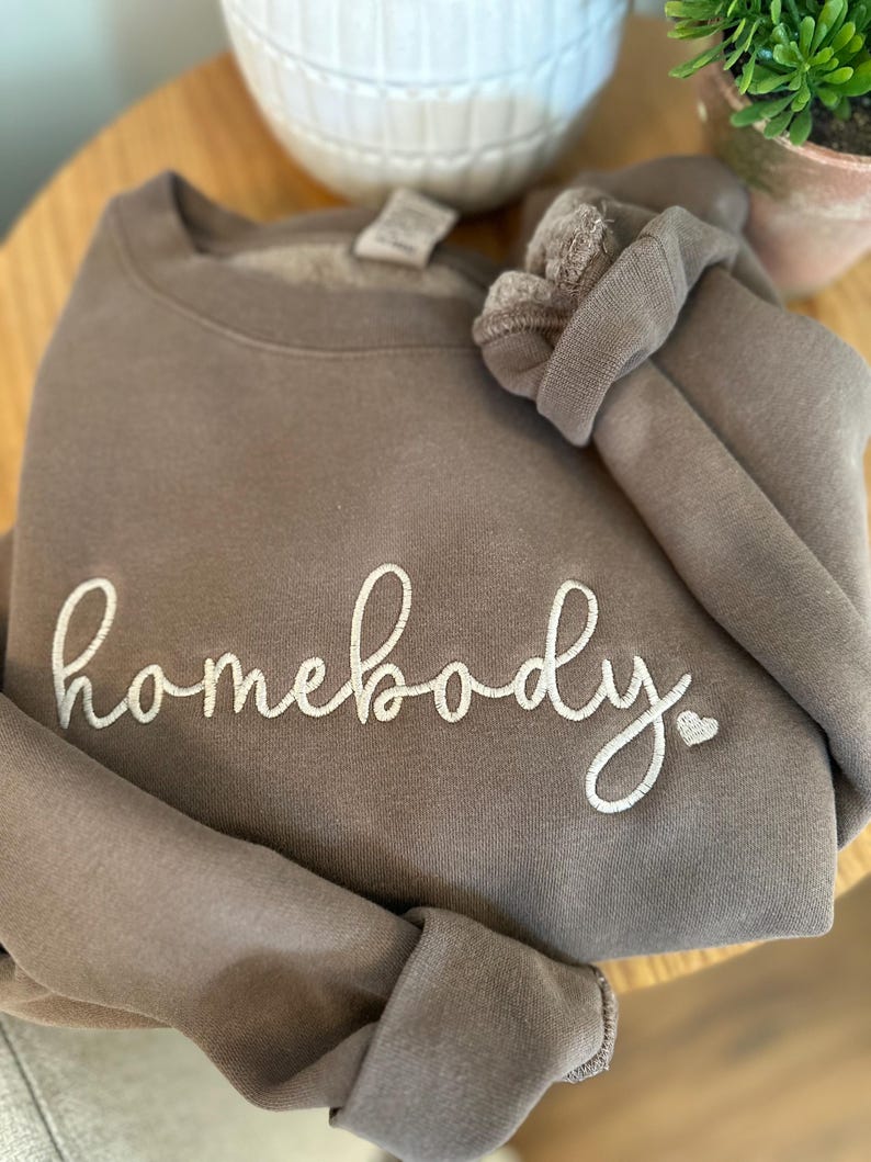 Peut inclure: Un sweat-shirt marron avec le texte "homebody" brod&eacute; en blanc avec un petit c&oelig;ur.