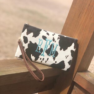 Puede incluir: Un embrague de estampado de vaca blanco y negro con una correa de mu&ntilde;eca marr&oacute;n. El embrague tiene un monograma turquesa "RFM" bordado en la parte delantera.