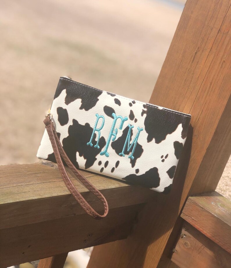 Puede incluir: Un embrague de estampado de vaca blanco y negro con una correa de mu&ntilde;eca marr&oacute;n. El embrague tiene un monograma turquesa "RFM" bordado en la parte delantera.