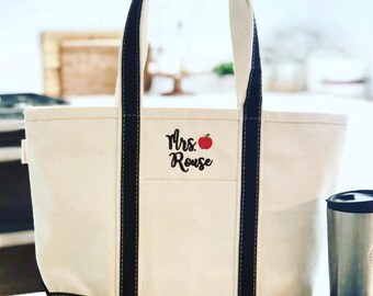 May include: A white canvas tote bag with black handles and trim. The bag has an embroidered design that says "Mrs. Rouse" with a red apple.