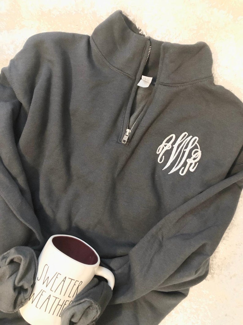May include: Dark gray quarter-zip sweatshirt with a white monogram on the chest. A white mug with the words "SWEATER WEATHER" is visible. The mug has a dark red interior.