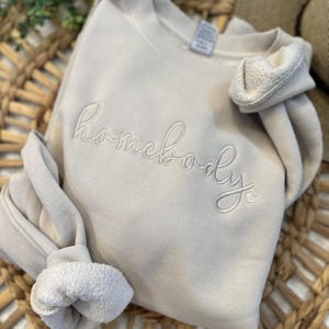 Peut inclure: Un sweat-shirt beige clair avec le mot "homebody" brod&eacute; en blanc sur le devant.