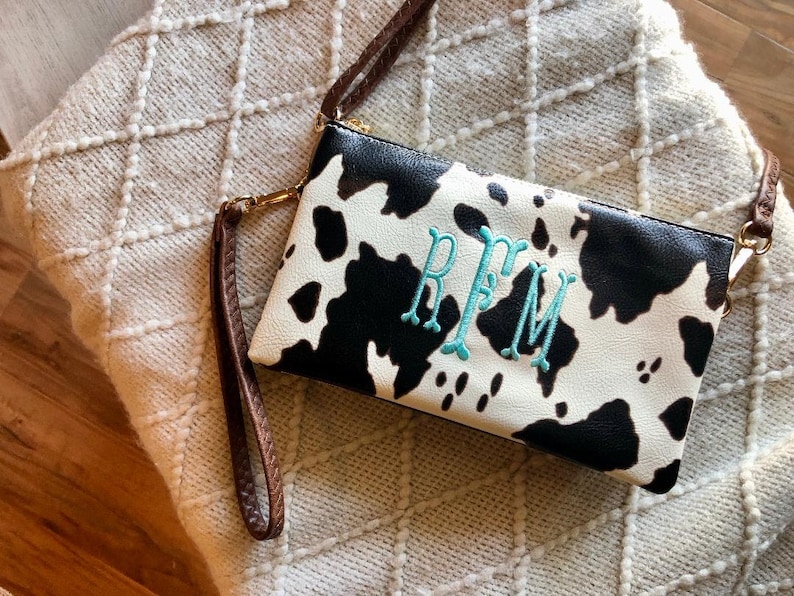 Puede incluir: Un bolso de mano de estampado de vaca en blanco y negro con una correa marr&oacute;n. El bolso tiene un monograma turquesa "RFM" bordado en la parte delantera.