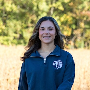 May include: Navy blue quarter-zip sweatshirt with a white monogram on the left chest. The sweatshirt has long sleeves and a zipper closure. The background is a blurred view of a field and trees.