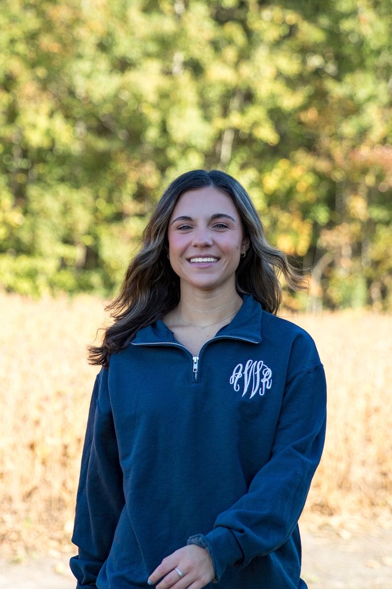 May include: Navy blue quarter-zip sweatshirt with a white monogram on the left chest. The sweatshirt has long sleeves and a zipper closure. The background is a blurred view of a field and trees.