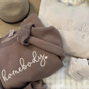 Peut inclure: Deux sweats &agrave; capuche avec le texte "homebody" brod&eacute; dessus. Le sweat &agrave; capuche de gauche est marron avec un c&oelig;ur blanc &agrave; c&ocirc;t&eacute; du texte. Le sweat &agrave; capuche de droite est beige clair avec le texte en blanc.