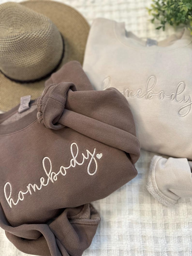 Peut inclure: Deux sweats &agrave; capuche avec le texte "homebody" brod&eacute; dessus. Le sweat &agrave; capuche de gauche est marron avec un c&oelig;ur blanc &agrave; c&ocirc;t&eacute; du texte. Le sweat &agrave; capuche de droite est beige clair avec le texte en blanc.
