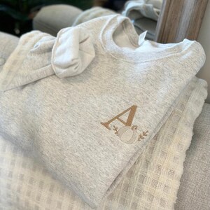 May include: A grey sweatshirt with a brown embroidered letter "A" and a pumpkin design. The sweatshirt is folded and placed on top of a white blanket.