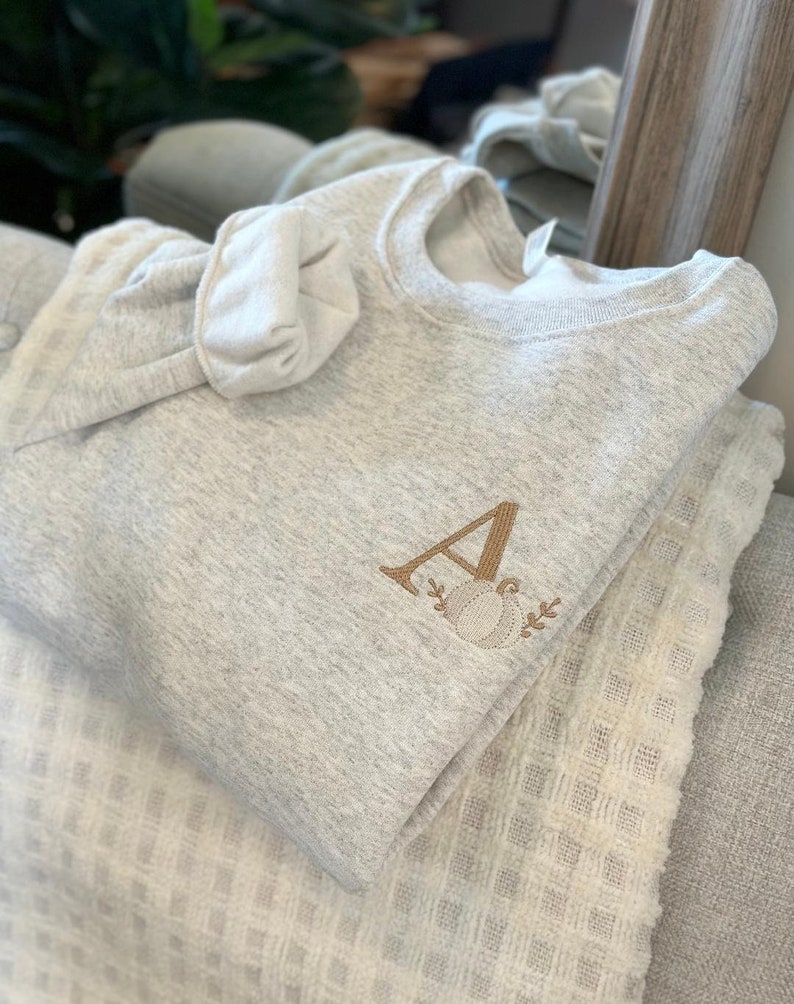 May include: A grey sweatshirt with a brown embroidered letter "A" and a pumpkin design. The sweatshirt is folded and placed on top of a white blanket.