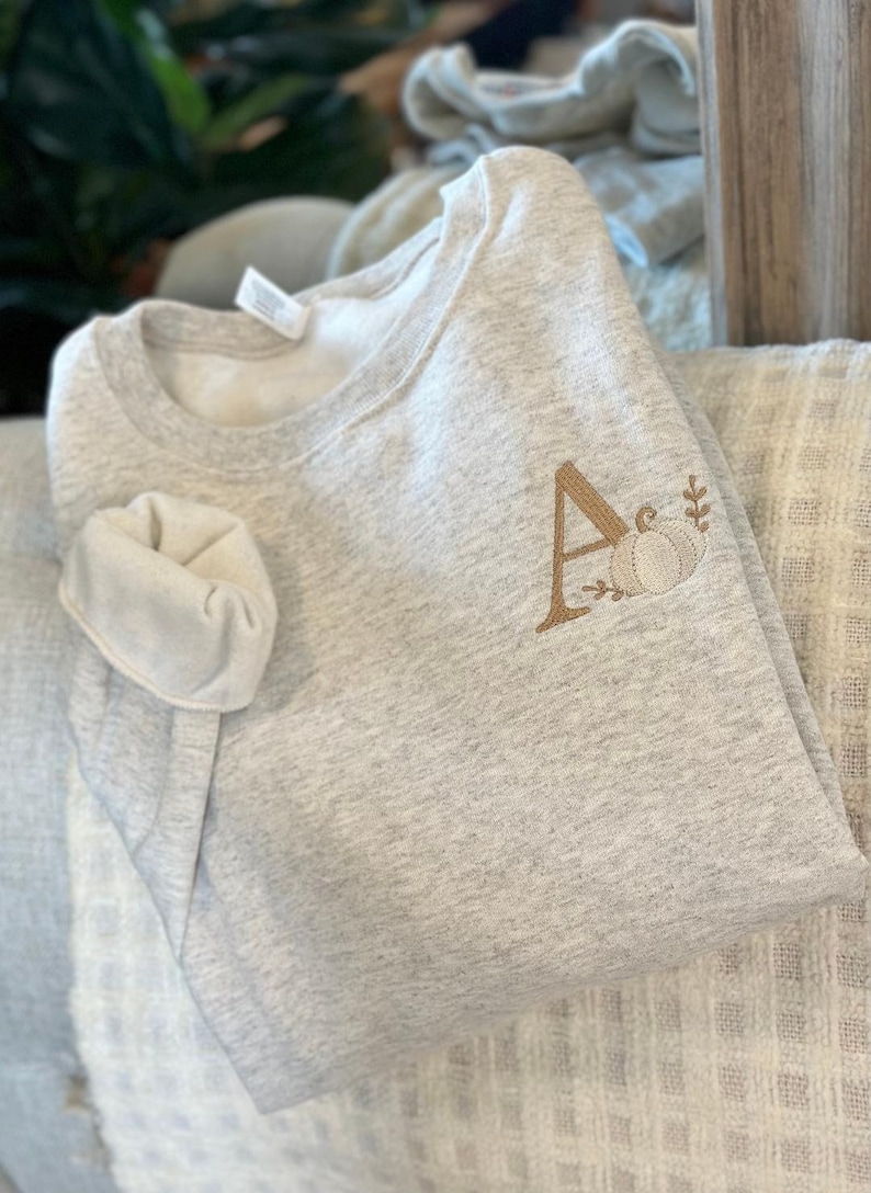 May include: A grey sweatshirt with a brown embroidered letter A and a pumpkin design.
