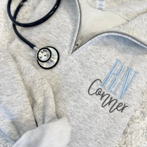 May include: A light gray quarter-zip sweatshirt with the embroidered letters "RN" in light blue and the name "Conner" in gray. A black stethoscope rests on the sweatshirt. The sweatshirt has a silver zipper.